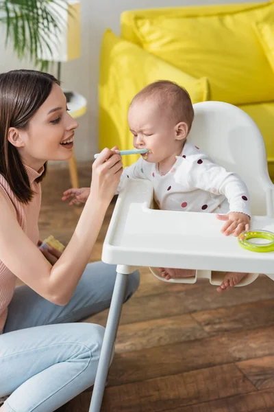 Happy woman feeding little son sitting in baby chair with closed eyes — Stock Photo
