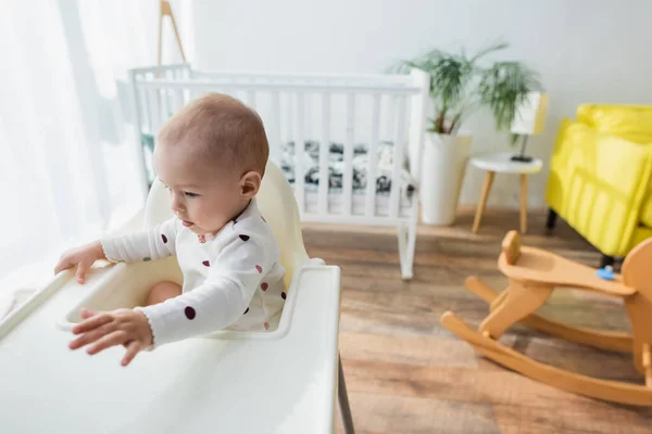 Tout-petit garçon assis dans une chaise bébé près de la crèche floue et cheval à bascule — Photo de stock