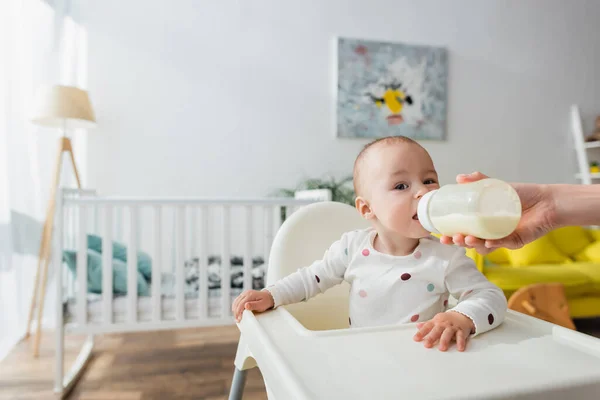 Femme avec du lait dans un biberon nourrissant son fils près d'un berceau flou — Photo de stock