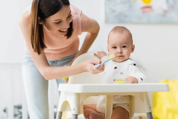 Little boy in romper sitting in baby chair while happy mother feeding him from spoon — Stock Photo
