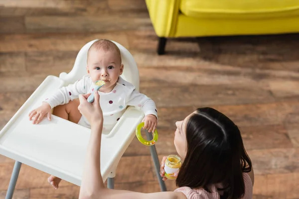 Bambino ragazzo guardando fotocamera mentre la madre lo alimenta con purea a casa — Foto stock