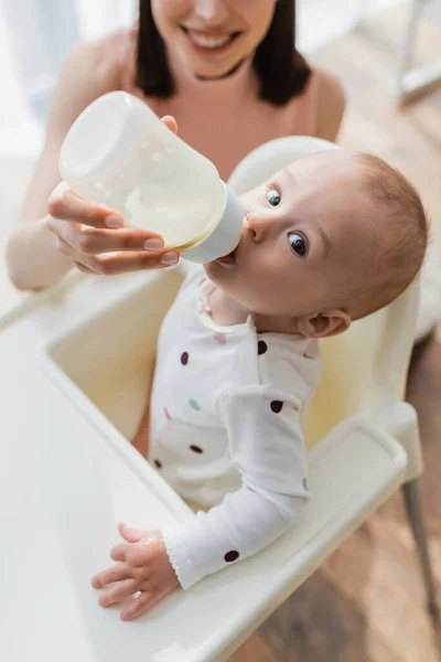 Hochwinkelaufnahme eines kleinen Kindes, das in die Kamera schaut, während es Milch aus einer Babyflasche in der Hand einer verschwommenen Mutter trinkt — Stockfoto