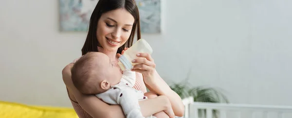 Щаслива жінка з пляшкою з немовлям годує маленького сина вдома, банер — стокове фото