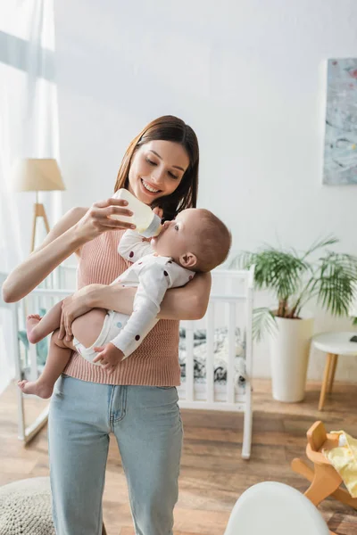 Glückliche Frau füttert kleinen Sohn mit Milch, während sie in der Nähe der verschwommenen Krippe steht — Stockfoto