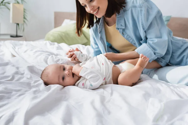 Tout-petit enfant tenant la main dans la bouche tout en étant couché sur le lit près de la mère gaie — Photo de stock