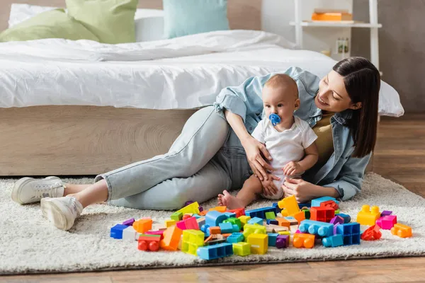 Baby boy with pacifier and his happy mom playing with colorful building blocks on floor — Stock Photo
