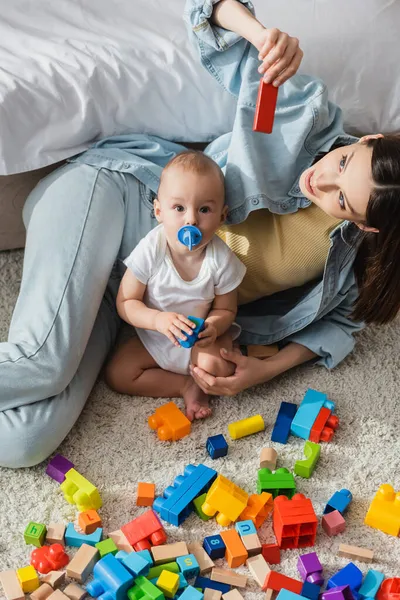 High angle view of little boy looking at camera near mother and building blocks on floor — Stock Photo