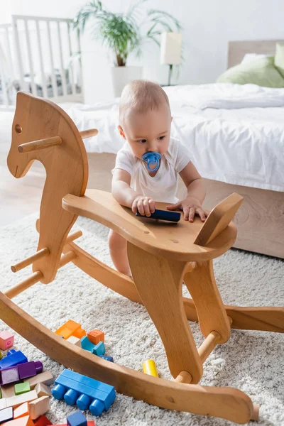 Toddler kid with pacifier standing near rocking horse and building blocks on floor — Stock Photo