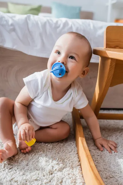 Cheerful little boy with pacifier holding toy building block while sitting on floor — Stock Photo