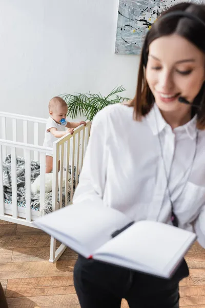 Verschwommene Frau mit Kopfhörer hält leeres Notizbuch, während sie in der Nähe ihres Sohnes im Kinderbett zu Hause arbeitet — Stockfoto