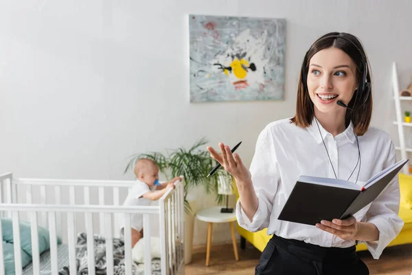 Smiling woman talking in headset near baby boy in crib on blurred background — Stock Photo
