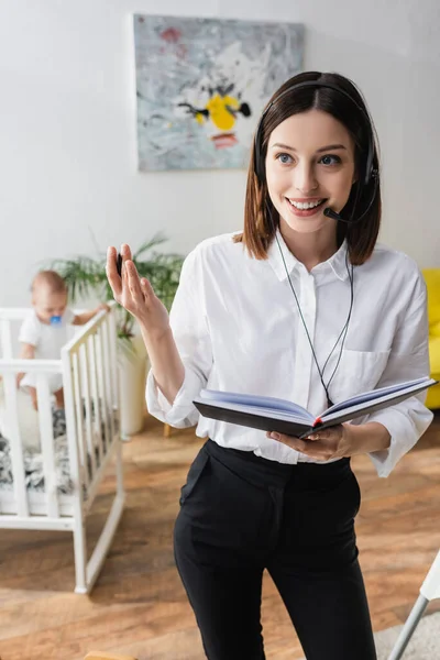 Happy woman in headset holding notebook while working near little son in crib on blurred background — Stock Photo