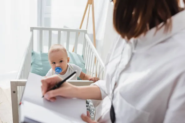 Verschwommene Frau schreibt in Notizbuch, während sie nahe Kind in Krippe arbeitet — Stockfoto