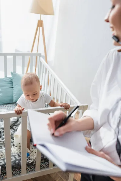 Mujer borrosa escribiendo en un cuaderno cerca de un niño pequeño en una cuna - foto de stock