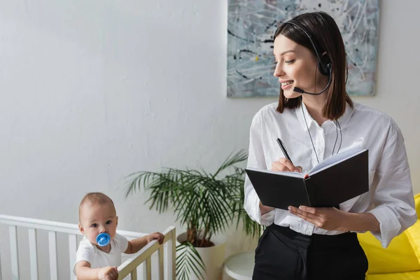 Frau mit Headset schreibt in Notizbuch, während sie zu Hause in der Nähe von Baby im Kinderbett arbeitet — Stockfoto