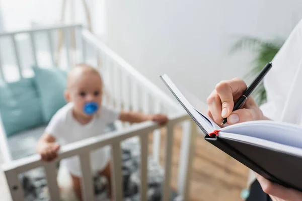 Woman writing in notebook near blurred baby boy in crib — Stock Photo