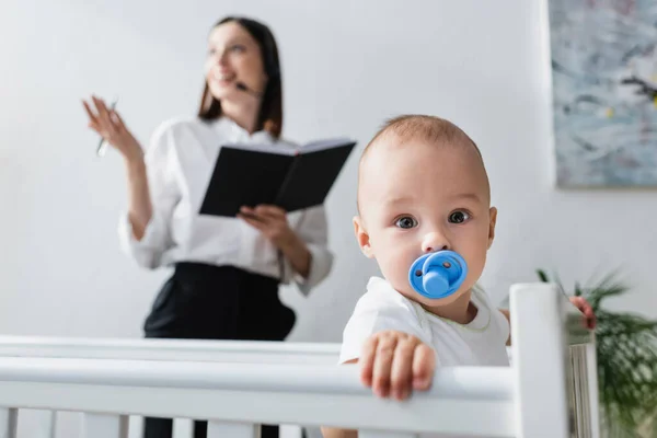 Baby boy with pacifier looking at camera near blurred mother working at home — Stock Photo