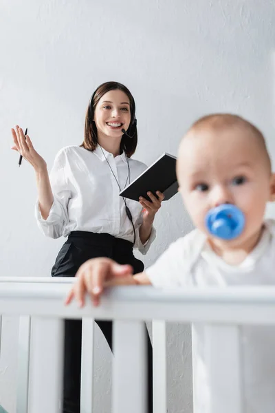 Mujer feliz con cuaderno de trabajo en auriculares cerca borrosa niño pequeño en cuna - foto de stock