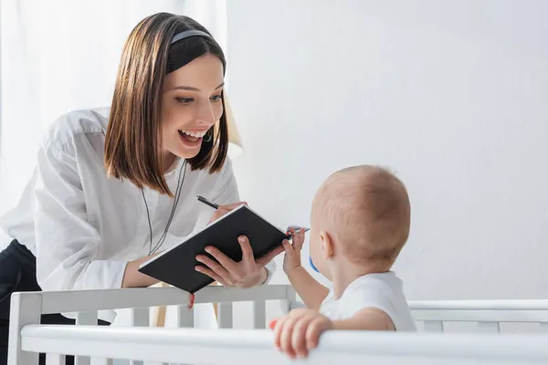 Happy woman in headset writing in notebook near toddler son in crib — Stock Photo