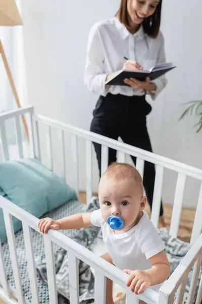 Baby boy looking at camera from crib near mom working on blurred background — Stock Photo