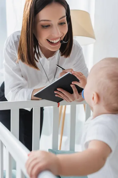 Alegre mujer en auriculares escritura en cuaderno cerca borrosa hijo en cuna - foto de stock