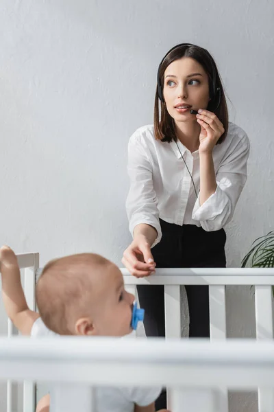 Blurred boy in crib near mother working in headset at home — Stock Photo