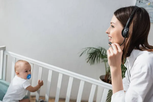 Glückliche Frau spricht in Kopfhörer neben verschwommenem Kleinkind im Kinderbett — Stockfoto