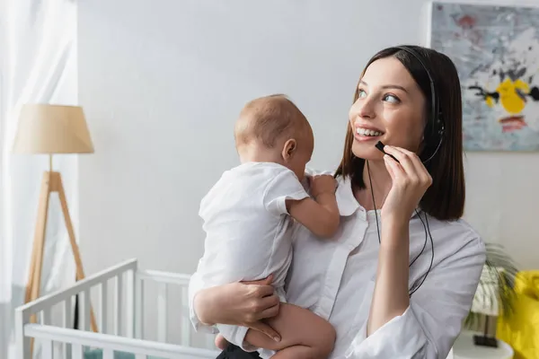 Улыбающаяся женщина в наушниках держит малыша сына во время работы дома — стоковое фото