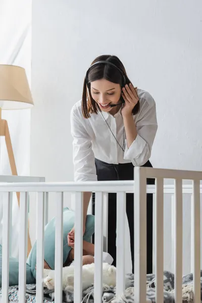 Mujer positiva hablando en auriculares cerca de la cuna con el niño pequeño - foto de stock