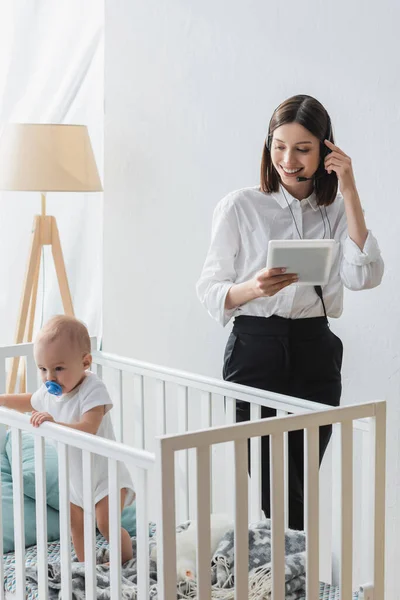 Smiling woman with digital tablet working in headset near toddler child in crib — Stock Photo