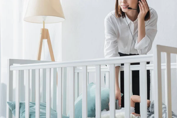 Cropped view of woman in headset working near baby crib at home — Stock Photo