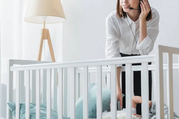 Vista recortada de la mujer en auriculares trabajando cerca de la cuna en casa - foto de stock