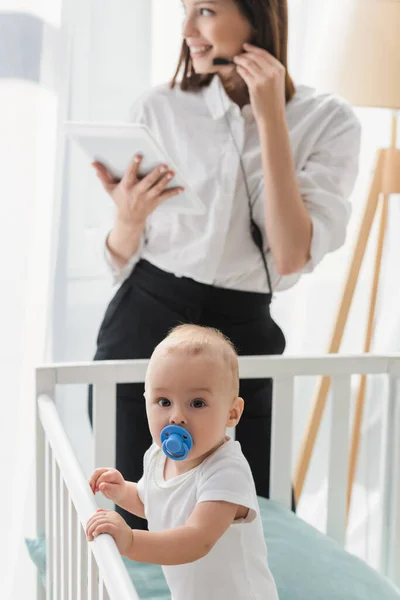 Junge in Krippe blickt in die Kamera nahe der Mutter mit digitalem Tablet, das auf verschwommenem Hintergrund arbeitet — Stockfoto