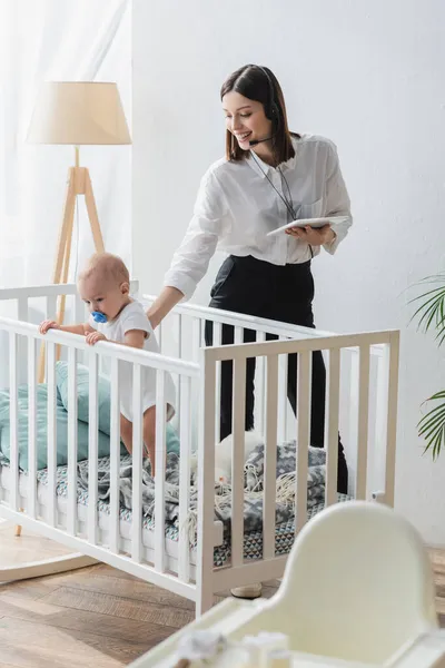 Frau mit Headset hält digitales Tablet in der Hand, während sie neben kleinem Sohn im Kinderbett steht — Stockfoto