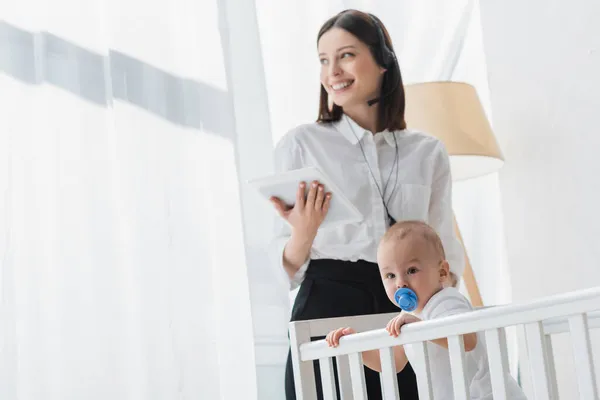 Smiling woman in headset holding digital tablet while standing near baby in crib — Stock Photo