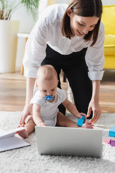 Donna bruna sorridente vicino al figlio del bambino seduto sul pavimento vicino al computer portatile offuscato — Foto stock