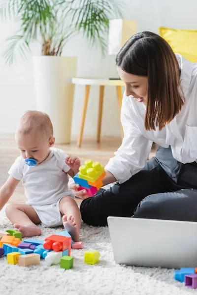 Bruna donna tenendo blocco di costruzione mentre seduto al piano vicino a piccolo figlio e laptop — Foto stock