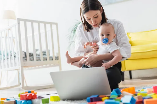 Madre sosteniendo niño pequeño mientras está sentado en el suelo cerca de la computadora portátil y borrosa bloques de construcción - foto de stock