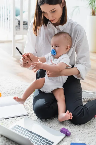 Femme montrant stylo au petit fils tout en étant assis sur le sol près d'un ordinateur portable — Photo de stock