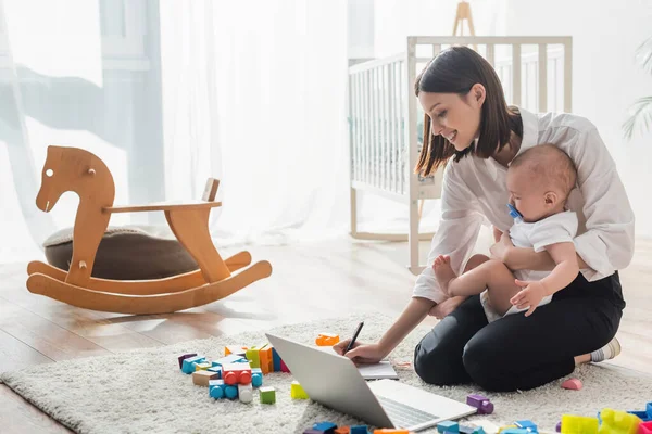 Mujer sonriente escribiendo en un cuaderno mientras está sentada en el suelo con su hijo cerca de la computadora portátil y bloques de construcción - foto de stock