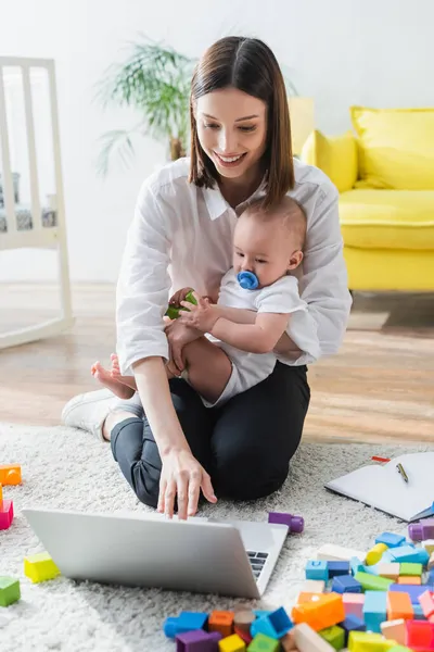 Lächelnde Frau mit Laptop, während sie mit Sohn in der Nähe von Bauklötzen auf dem Boden sitzt — Stockfoto