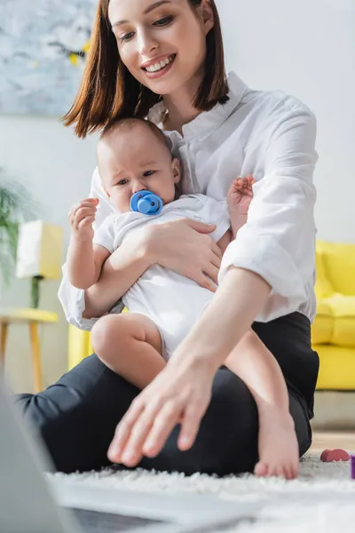 Smiling woman holding little son while sitting on floor near blurred laptop — Stock Photo