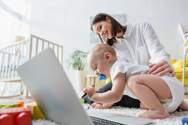 Малыш держит ручку рядом с счастливой мамой и ноутбуком на полу — стоковое фото
