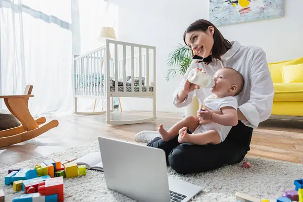 Donna sorridente che parla sullo smartphone mentre alimenta il figlio al piano vicino a laptop e blocchi di costruzione — Foto stock