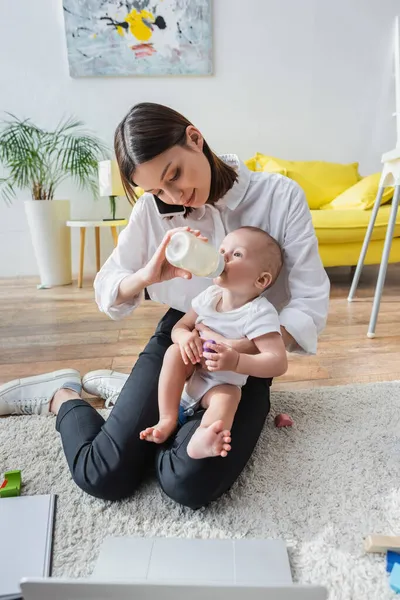 Mulher morena falando no celular enquanto alimenta o filho com leite no chão perto do laptop — Stock Photo