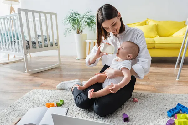 Mujer feliz sentada en el suelo cerca de la computadora portátil y alimentando a su pequeño hijo del biberón - foto de stock