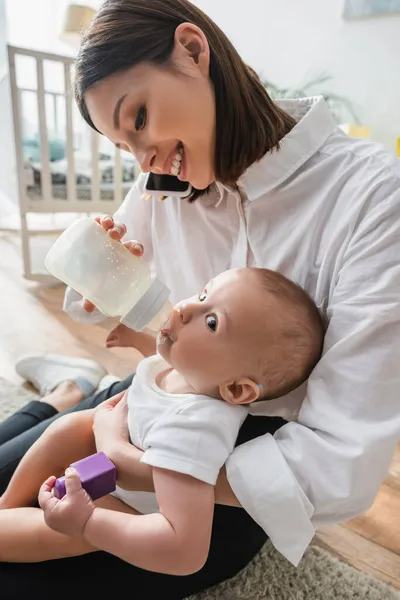 Smiling woman talking on mobile phone while feeding little son with milk at home — Stock Photo