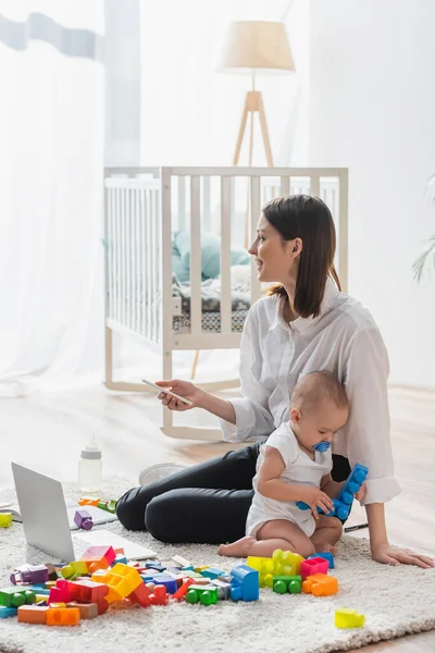 Glückliche Frau mit Handy sitzt auf dem Boden neben Kleinkind Sohn spielt mit Bauklötzen — Stockfoto