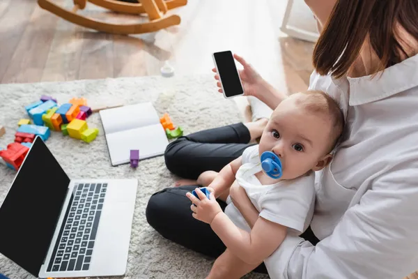 Mujer con teléfono inteligente sentado en el suelo con bebé niño cerca de la computadora portátil y bloques de construcción - foto de stock