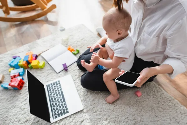 Vista de ángulo alto de la mujer con su hijo pequeño y teléfono inteligente sentado en el suelo cerca de la computadora portátil y bloques de construcción - foto de stock